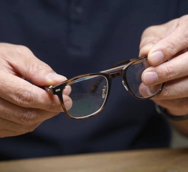 A close-up of a pair of handcrafted glasses being polished by an artisan, highlighting the detail and quality expected of the profession. Neutral palette with dark navy backgrounds. North American / US / Indiana.
