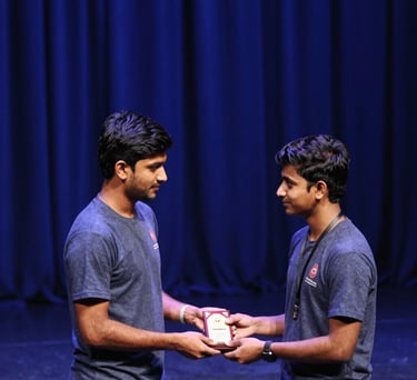 A high-angle shot of a South Asian student athlete receiving an award on a stage. Royal blue curtains in the background and soft stage lighting.