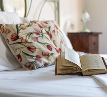 An open hardcover book rests on a white bed with floral pillows in a cozy bedroom setting.