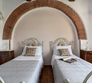 Rustic twin bedroom with ornate metal headboards under a large brick archway in an Italian villa.