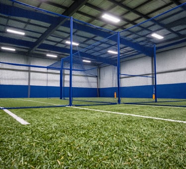 A low-angle shot of a high-performance softball training facility in the North American / US Midwest, showing indoor hitting tunnels with blue netting and professional turf, under bright, clean white lighting.