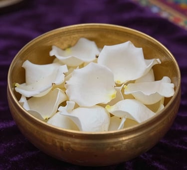 A close-up photograph of a polished gold bowl filled with fresh white rose petals, set on a dark purple velvet cloth in a South American spiritual sanctuary. Soft ambient lighting.
