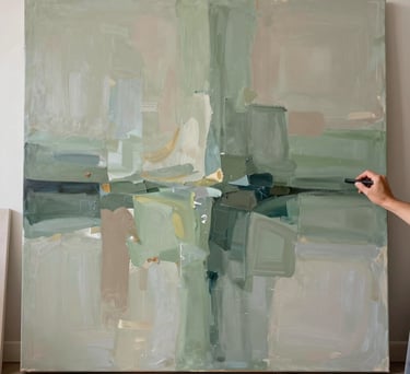 Photography of a large abstract painting in progress, with soft washes of sage green and taupe. The artist's hand is visible in the corner, holding a charcoal stick, in a sun-drenched North American studio.