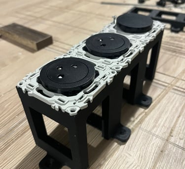 Three-gang black electrical socket outlets mounted on a custom structural bracket on a wooden workbench.