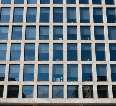 Photography of an official building facade with modern glass architecture reflecting a blue sky. Clean, authoritative, and professional. Medium blue and off-white tones. Global / Spanish-speaking.