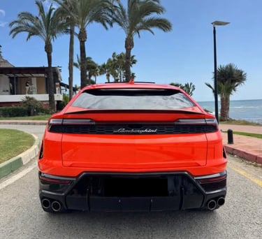 Rear view of a bright orange Lamborghini Urus luxury SUV parked by palm trees near the ocean.