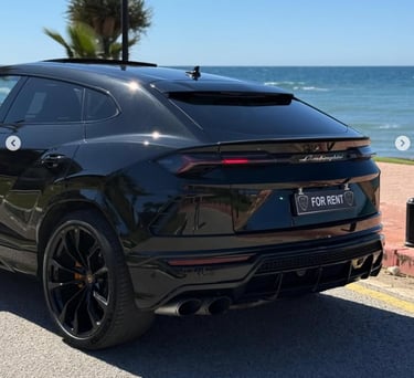Black Lamborghini Urus luxury SUV available for rent parked on a coastal road by the ocean.