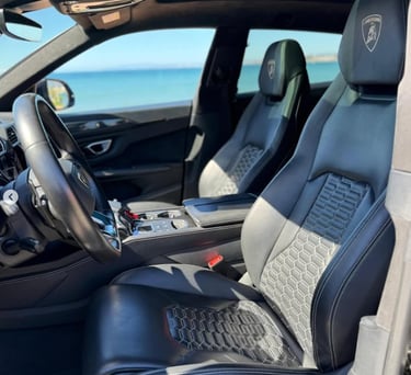 Luxury interior of a Lamborghini Urus with black leather seats and honeycomb stitching parked by the ocean.