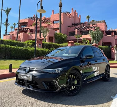 Black Volkswagen Golf R for rent parked in front of a luxury pink resort with palm trees.