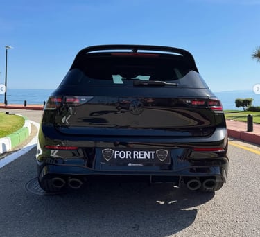 Rear view of a black Volkswagen Golf R for rent parked on a coastal road by the sea.