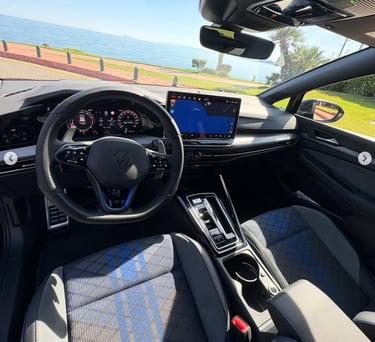 Interior view of a modern Volkswagen Golf with blue accent seats and digital cockpit dashboard parked by the sea.