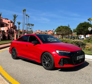 A vibrant red Audi RS3 luxury sedan parked on a sunny resort road with palm trees and blue sky.