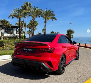 A bright red Audi RS3 luxury rental car parked on a coastal road with palm trees and ocean views.