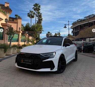 White Audi RS3 luxury hatchback parked on a paved street with palm trees and Mediterranean architecture in the background.