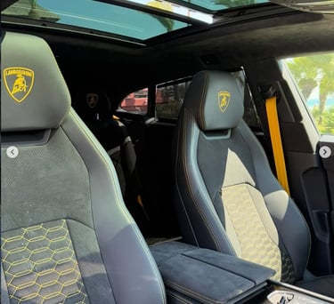Interior of a luxury Lamborghini Urus featuring black leather seats with yellow stitching and panoramic sunroof.