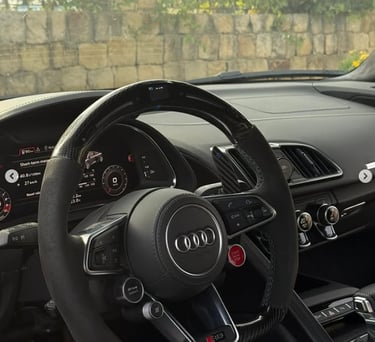 Interior view of an Audi R8 luxury sports car showing the carbon fiber steering wheel and digital dashboard.