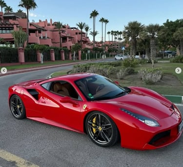 A red Ferrari 488 luxury sports car parked on a coastal road lined with palm trees and pink buildings.
