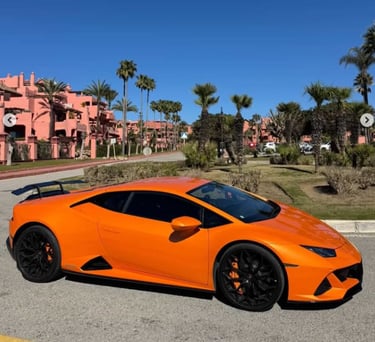 Orange Lamborghini Huracan supercar with black wheels parked near tropical palm trees and pink buildings.