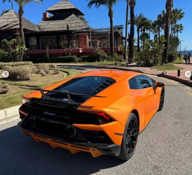 Orange Lamborghini Huracan supercar parked on a sunny road near a tropical resort with palm trees.