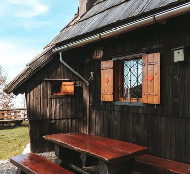 Rustic wooden alpine cabin with heart-carved shutters and an outdoor picnic table on a stone patio.