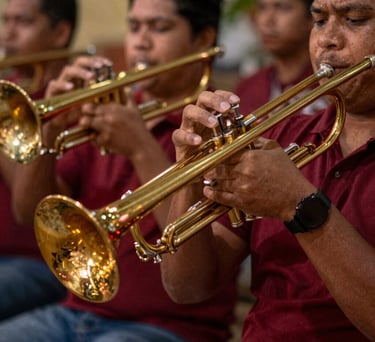 A close-up South American / Brazilian Amazonian musical photography shot. Dynamic, warm-lit capture of hands playing brass instruments like trombones and trumpets, reflecting gold and deep carmine red festive lights.