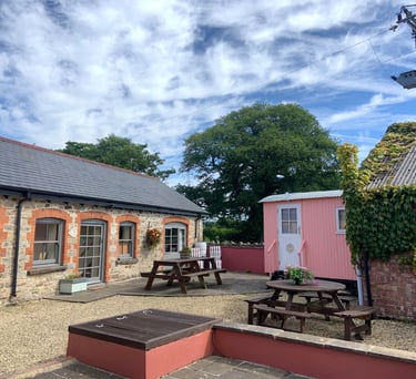 Rustic farm courtyard with a stone cottage, pink shepherd hut, and wooden picnic tables under a blue sky.
