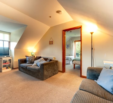 Cozy attic living room with grey corduroy sofas, beige carpet, and a view into a bright bedroom.