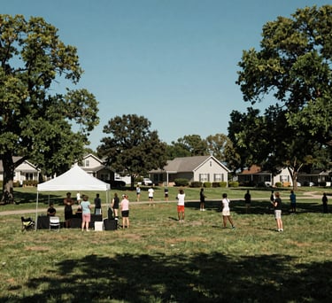 A wide-angle photography shot of a North American suburban community park where a wellness event is taking place. People are gathered under a white tent, with lush green trees and a clear blue sky. The scene is filled with a sense of stability and peace.
