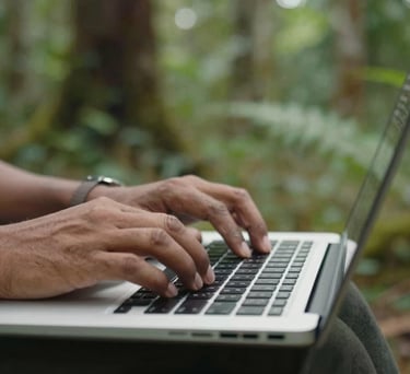 Close-up of a data specialist's hands using a laptop in the field, with an Indonesian forest in the blurred background, representing data-driven conservation.