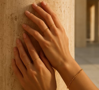 Close-up editorial photograph of a woman's hands wearing delicate gold minimalist rings and a classic chain bracelet, gently touching a warm sand-toned travertine stone column. Soft, warm natural light, high-end fashion catalog style, set in a modern Western European open-air space.