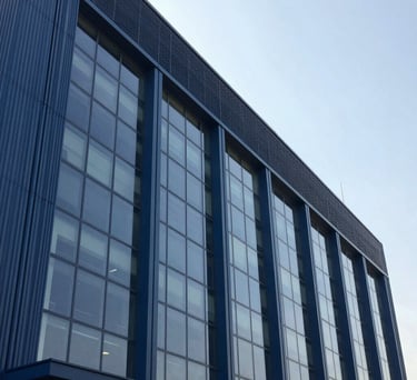 A photography shot of the exterior of a sleek, modern industrial building in a Pakistani business district, characterized by large glass windows and steel blue architectural panels. The sky is clear and bright.
