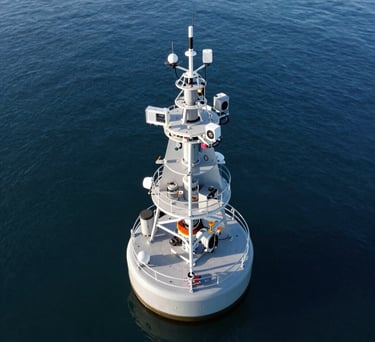 An aerial view of an environmental monitoring project in a North American / Global coastal area. High-tech sensors are visible on a modern buoy, set against deep blue water and grey equipment.