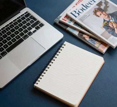 An overhead shot of a laptop, a notebook, and a scattered collection of literary magazines on a dark blue surface, sophisticated lighting.
