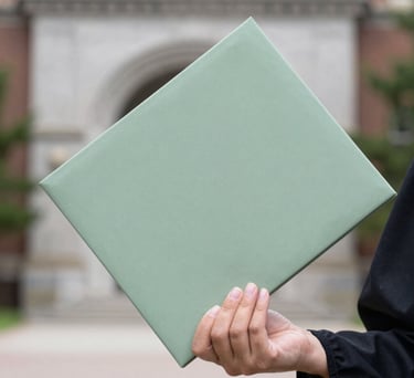 A close-up photograph of a graduate's hand holding a diploma folder, symbolizing academic success and opportunity, with a soft-focus North American university background in light gray and sage green tones.