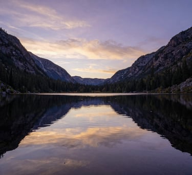 A wide-angle landscape photograph of a calm, reflective pond in a North American mountain valley. The water is still, mirroring a sky tinged with soft purple and gold at sunrise. The composition is peaceful and vast.