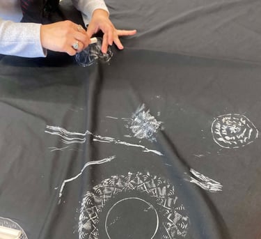 A young person using a stick of chalk on fabric to rub the outlines of a shape underneath the fabric.