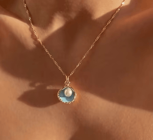 A simple gold necklace with half a shell as a pendant. A pearl hangs from the chain to sit in it.