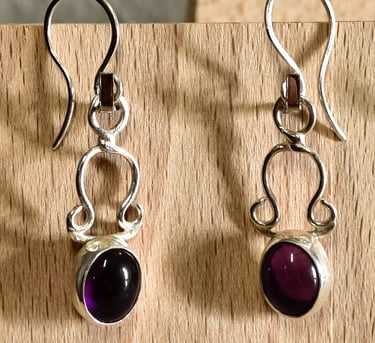 Handmade sterling silver drop earrings with oval purple amethyst gemstones on a wooden background.