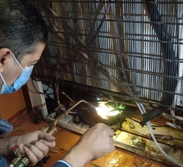A technician wearing a mask uses a welding torch to repair a refrigerator compressor and cooling coils.