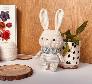 a small handmade amigurumi white bunny, with body in stripes, with a potted plant in the background