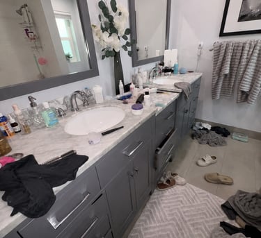 Messy modern bathroom with dual gray vanities, marble countertops, and clothes on the floor.