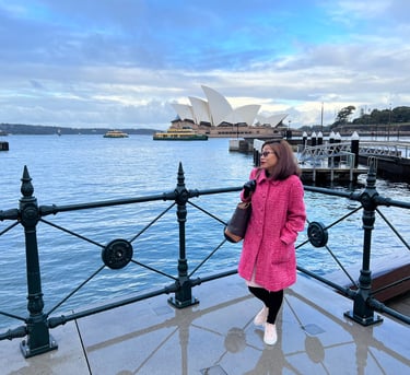 A woman in a pink coat stands on the foreground with the Sydney opera house in the background