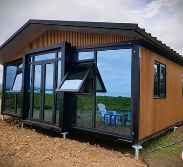 Modern prefab cabin with wood siding and large windows reflecting a green landscape.