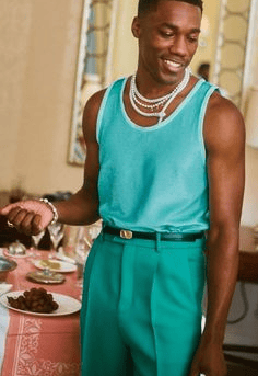Man wearing a teal tanktop and darker teal pants, with white shell necklaces and jewelry.