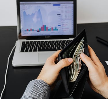 Notebook sobre uma mesa e duas mãos segurando uma carteira aberta com dinheiro dentro