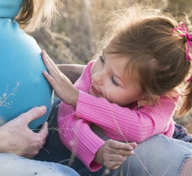 A young girl in a pink sweater leaning against her pregnant mother's belly in an outdoor field.