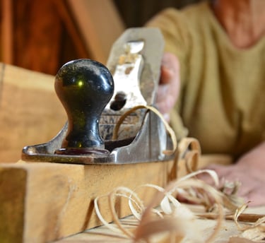 A woodworker uses a vintage metal hand plane to smooth a wooden board with curly shavings.