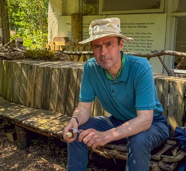 Simon is sitting on a wooden bench in the shade at Westbury Wildlife Park, whittling a mushroom.