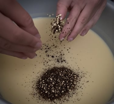 Hands sprinkling coffee grounds and oats into melted soap mixture