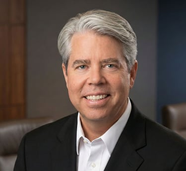 Professional headshot of a smiling male executive with grey hair in a black suit jacket.